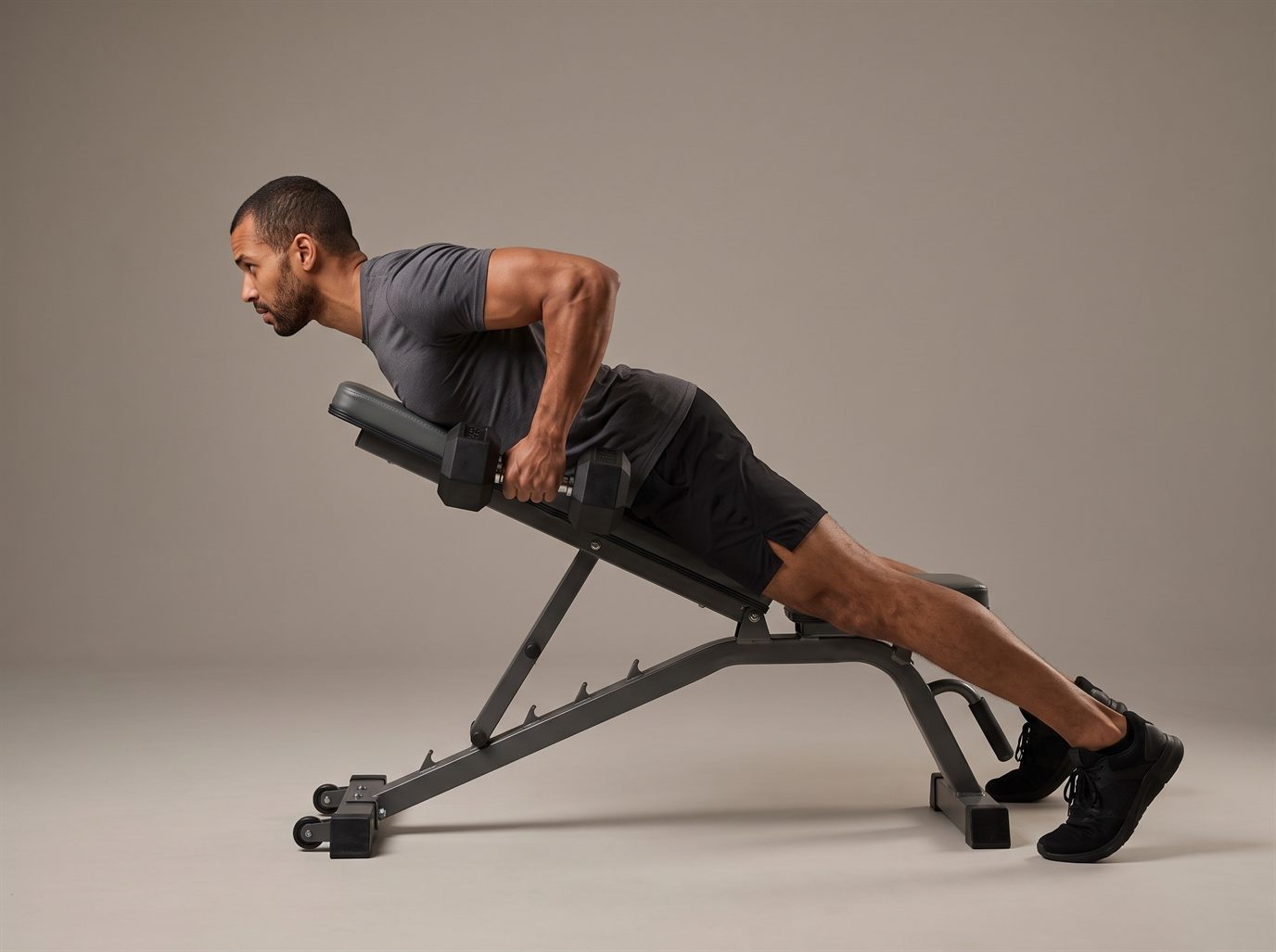 A man lying face down on an adjustable bench set to a low incline, rowing a pair of hex dumbbells with his elbows drawn close to his sides — a chest-supported row, one of the key dumbbell lat exercises.
