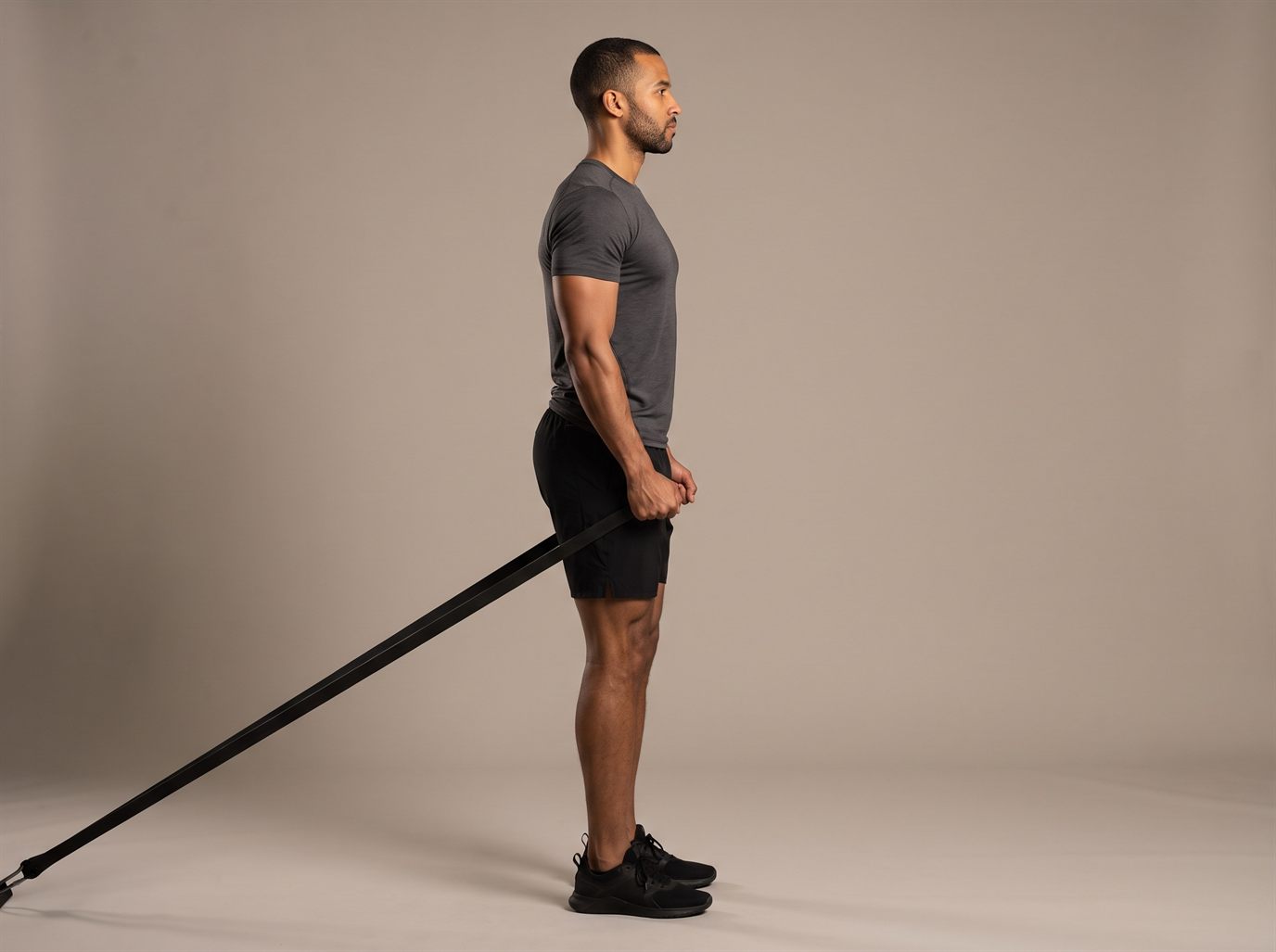 A man standing upright at the top of a band pull-through, hips fully extended and torso vertical, holding a resistance band that runs back between his legs to a low anchor behind him — a band-based hip thrust machine alternative.