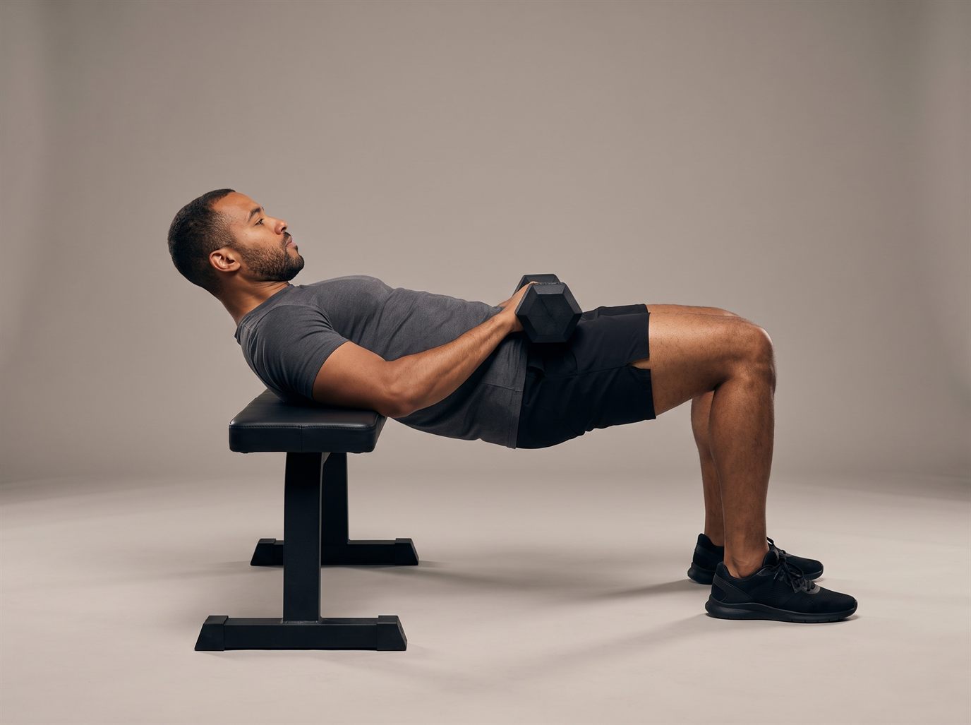 A man with his upper back on a flat bench and a hex dumbbell across his hips, driven up into the top of a dumbbell hip thrust with his shoulders, hips and knees level — the closest hip thrust machine alternative at home.