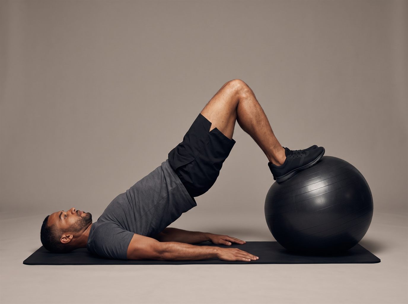 A man lying on his back on a mat with his heels on a black stability ball, hips lifted into a bridge and knees bent as he curls the ball toward him — a stability ball leg curl, the most practical leg curl alternative at home.