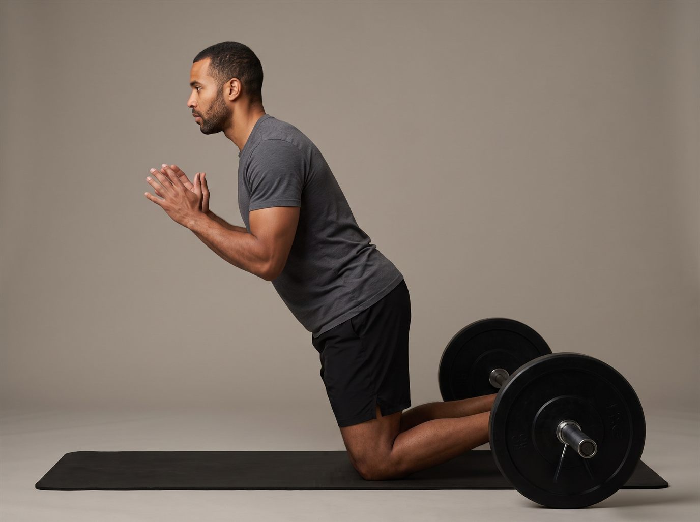 A man kneeling on a mat with his ankles pinned under a loaded barbell, leaning forward in one straight line from knees to shoulders with his hands raised, ready to catch himself — the Nordic ham curl, a leg curl alternative with no machine.