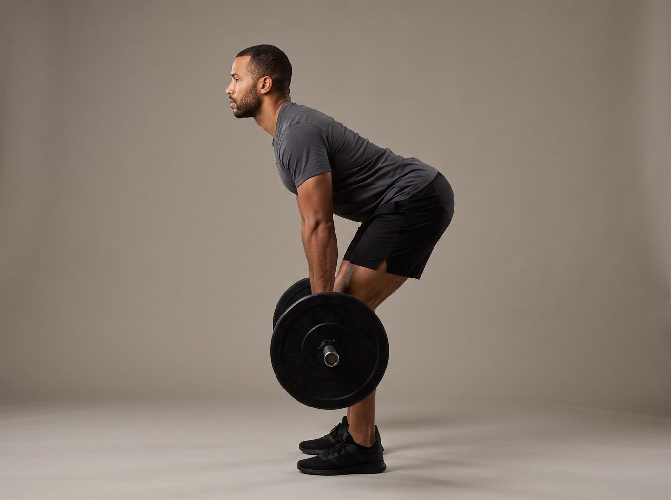 A man standing side-on in a Romanian deadlift, hips pushed back, back flat and knees slightly bent, holding a loaded barbell just below his knees — the hip-hinge exercise to pair with a leg curl alternative.
