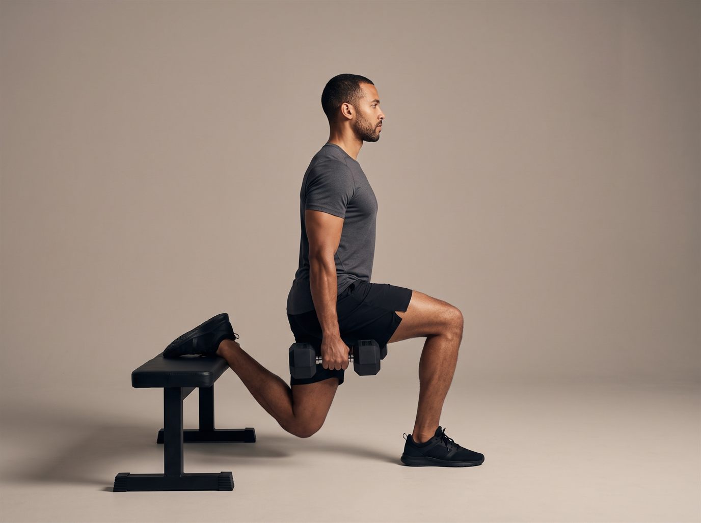 A man at the bottom of a Bulgarian split squat, rear foot resting laces-down on a flat bench, front thigh parallel to the floor and a dumbbell in each hand — the main leg press alternative for home.