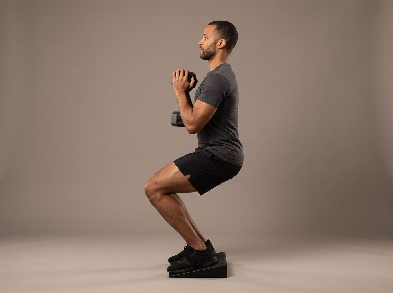 A man with his heels raised on a black wedge, holding one dumbbell vertically against his chest and squatting with his knees forward and torso upright — a heel-elevated goblet squat used as a leg press alternative.