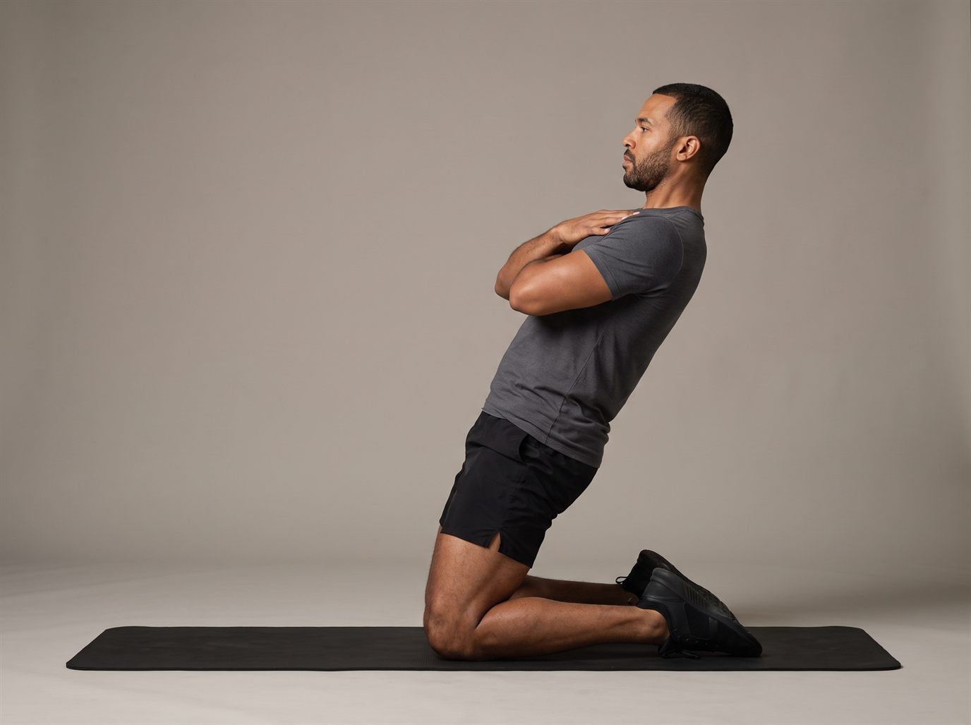 A man kneeling on a mat with his arms crossed over his chest, leaning back about 25 degrees with his knees, hips and shoulders in one straight line — a reverse Nordic, a bodyweight quad exercise to pair with a leg press alternative.