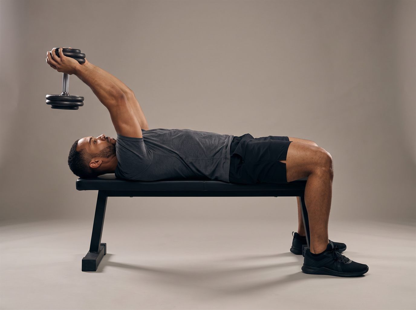 A man lying on a flat bench holding one dumbbell with both hands, arms extended overhead and slightly behind his head at the bottom of a dumbbell pullover, feet flat on the floor.