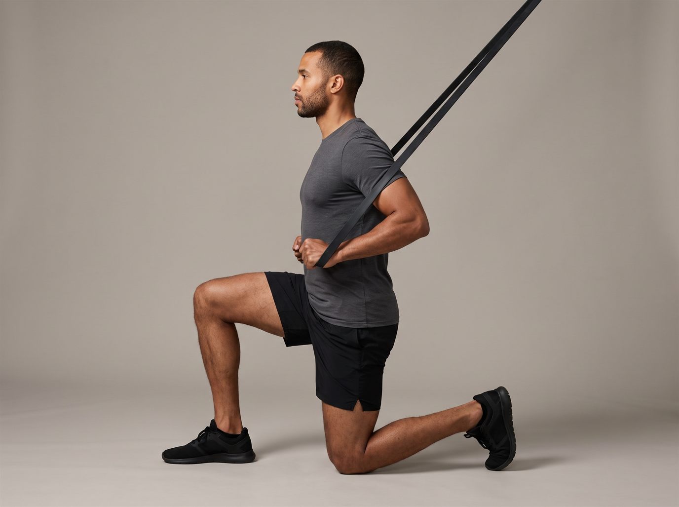 A man in a grey T-shirt half-kneeling at the finish of a band lat pulldown alternative, elbows drawn down beside his ribs, torso upright, the band running up and away to a high anchor.