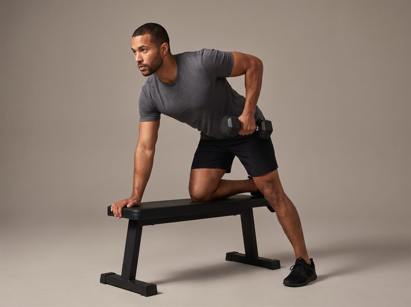 A man bracing one hand and one knee on a flat bench, torso angled forward, rowing a hex dumbbell up toward his hip as a lat-biased one-arm row.